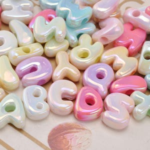 May include: A collection of assorted alphabet letter beads with a glossy, iridescent finish. The beads are rendered in pastel shades of pink, blue, yellow, and white, each featuring a central hole for stringing. The letters are shaped in a rounded, puffy style.