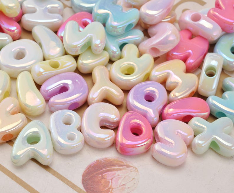 May include: A collection of assorted alphabet letter beads with a glossy, iridescent finish. The beads are rendered in pastel shades of pink, blue, yellow, and white, each featuring a central hole for stringing. The letters are shaped in a rounded, puffy style.