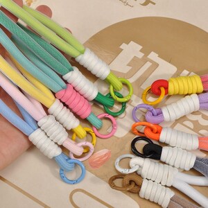May include: A collection of colorful wrist straps in various shades, including blue, pink, yellow, green, and purple. Each strap features a white wrapped section and a metal ring. The straps are designed for carrying small items.