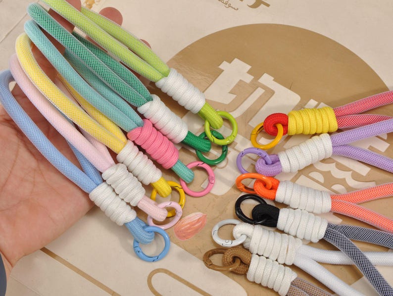 May include: A collection of colorful wrist straps in various shades, including blue, pink, yellow, green, and purple. Each strap features a white wrapped section and a metal ring. The straps are designed for carrying small items.