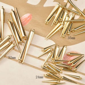May include: Gold-coloured metal cone-shaped jewellery findings in various sizes. The longest is 5.2cm, the shortest is 2.4cm, and another size is 3.5cm. Each cone has a loop at the top for attaching to other jewellery components. These findings are ideal for creating earrings, pendants, or other jewellery.