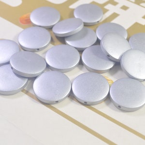 May include: A collection of silver-colored, flat, round beads with a matte finish. Each bead has a small hole. The beads are scattered across a light-colored surface, suggesting a craft or jewelry-making theme.