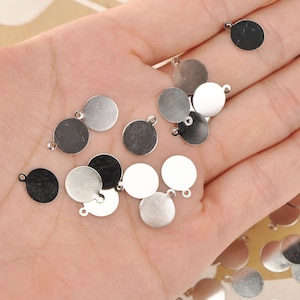 Puede incluir: Un conjunto de pequeños dijes redondos de metal plateado, cada uno con un lazo para sujetar a joyas u otras manualidades. Los dijes presentan varios tonos de plata y gris, con un acabado brillante.