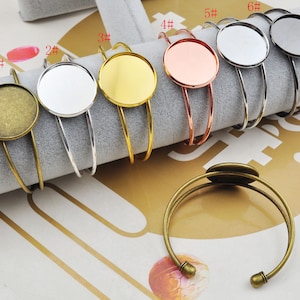 May include: Ten different styles of metal bangle bracelets with a round, flat, metal bezel for adding a cabochon. The bezels are in various colors, including gold, silver, copper, and black.