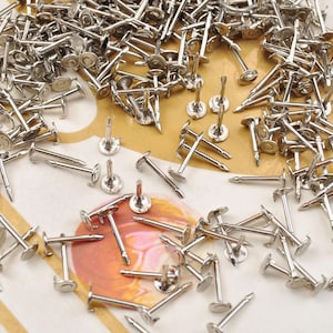 Op de afbeelding: Een grote hoeveelheid zilverkleurige metalen oorbelstiften met platte ronde achterkanten. De stiften zijn verspreid over een lichtbruin oppervlak.