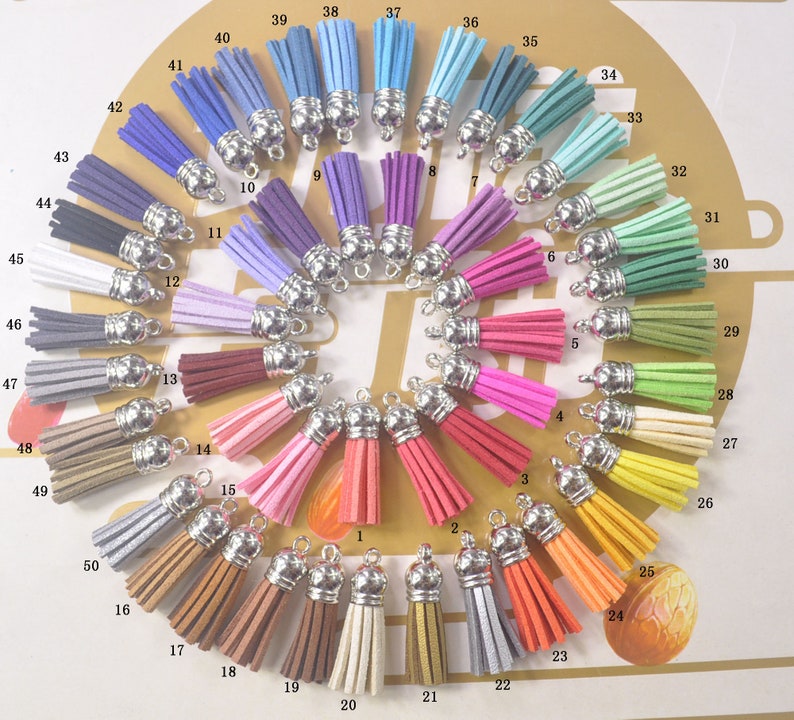 May include: A collection of 50 colorful suede tassels with silver-tone metal caps. The tassels are arranged in a circular pattern, showcasing a wide range of colors, including pinks, blues, greens, yellows, and browns. Each tassel is approximately 2 inches long.