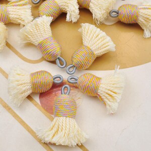 May include: A collection of cream-colored tassels with a multi-colored wrapped center and a silver-tone loop. The tassels have a fluffy, fringed bottom. The wrapped section features yellow, pink, and purple threads.
