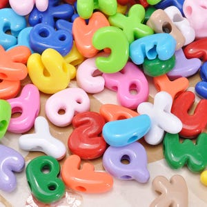 Op de afbeelding: Een verzameling kleurrijke plastic alfabetkralen in verschillende kleuren, waaronder roze, groen, oranje en blauw. De kralen hebben verschillende vormen, waaronder letters en cijfers, en hebben gaten om te rijgen. Ideaal voor knutselen en sieraden maken.