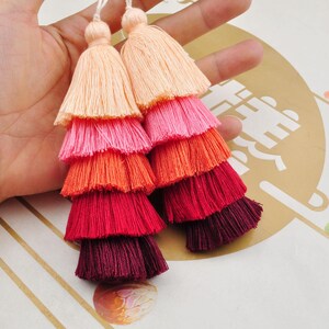 May include: Two multi-coloured tassel ornaments with a gradient of peach, pink, orange, red, and burgundy. The tassels are made of soft yarn and are layered to create a tiered effect.