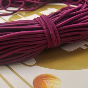 May include: A close-up of a large spool of deep burgundy elastic cord. The cord is tightly wound and has a smooth, shiny finish.