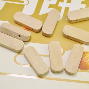 May include: A group of ten natural wood, unfinished, rectangular wooden beads with a hole drilled through the center of each bead. The beads are arranged on a light brown surface.