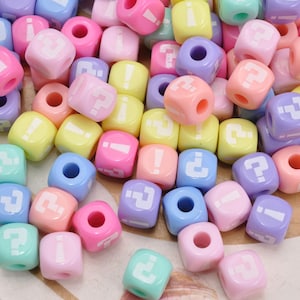 Op de afbeelding: Een verzameling kleine, kubusvormige plastic kralen in pastelkleuren zoals roze, geel, blauw, paars en mintgroen. Elke kraal is aan de voorzijde bedrukt met een wit vraagteken of uitroepteken en is voorzien van een gat in het midden.