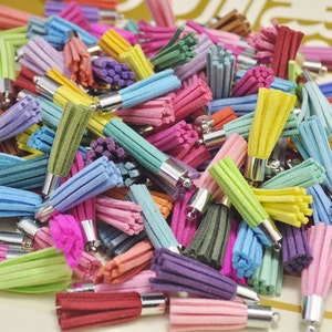 Op de afbeelding: Een grote verzameling kleurrijke suède kwastjes met zilveren metalen dopjes. De kwastjes zijn in een stapel gerangschikt en zijn in verschillende tinten blauw, groen, roze, paars, geel en rood.