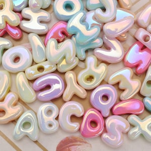 May include: A collection of assorted alphabet letter beads featuring a smooth, iridescent finish. The beads are rendered in a palette of pastel pink, blue, yellow, and lavender, each with a glossy, pearlescent sheen and a central hole for threading.