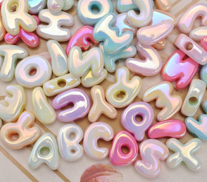May include: A collection of assorted alphabet letter beads featuring a smooth, iridescent finish. The beads are rendered in a palette of pastel pink, blue, yellow, and lavender, each with a glossy, pearlescent sheen and a central hole for threading.