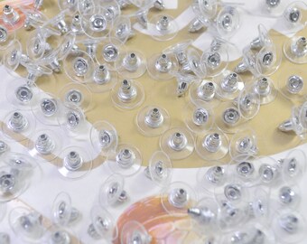 May include: A close-up of a large quantity of clear plastic earring backs with silver metal posts. The backs are scattered on a light brown surface.