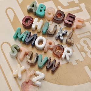 May include: A collection of colorful, translucent alphabet letters in various colors, including green, brown, white, and peach. The letters are arranged on a light brown patterned surface, creating a decorative display.