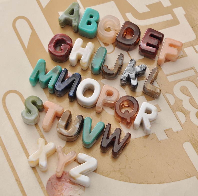 May include: A collection of colorful, translucent alphabet letters in various colors, including green, brown, white, and peach. The letters are arranged on a light brown patterned surface, creating a decorative display.