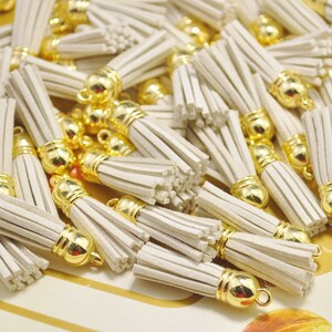 Könnte beinhalten: Ein Haufen cremefarbener Quasten mit goldenen Metallspitzen. Die Quasten bestehen aus einem weichen, flauschigen Material und sind perfekt, um jedem Projekt einen Hauch von Verspieltheit zu verleihen.