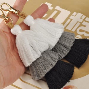 Peut inclure: Deux porte-clés à pompons superposés en blanc, gris et noir. Chaque pompon est attaché à un fermoir doré avec un cordon beige tissé. Les pompons sont faits de fils doux et superposés, créant un accessoire décoratif.
