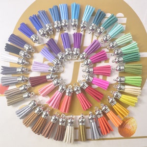 May include: A collection of colorful suede tassels in various shades, including blue, purple, pink, green, and orange. Each tassel has a silver-colored metal cap and a loop for attaching to jewelry or other crafts. The tassels are arranged in a circular pattern.