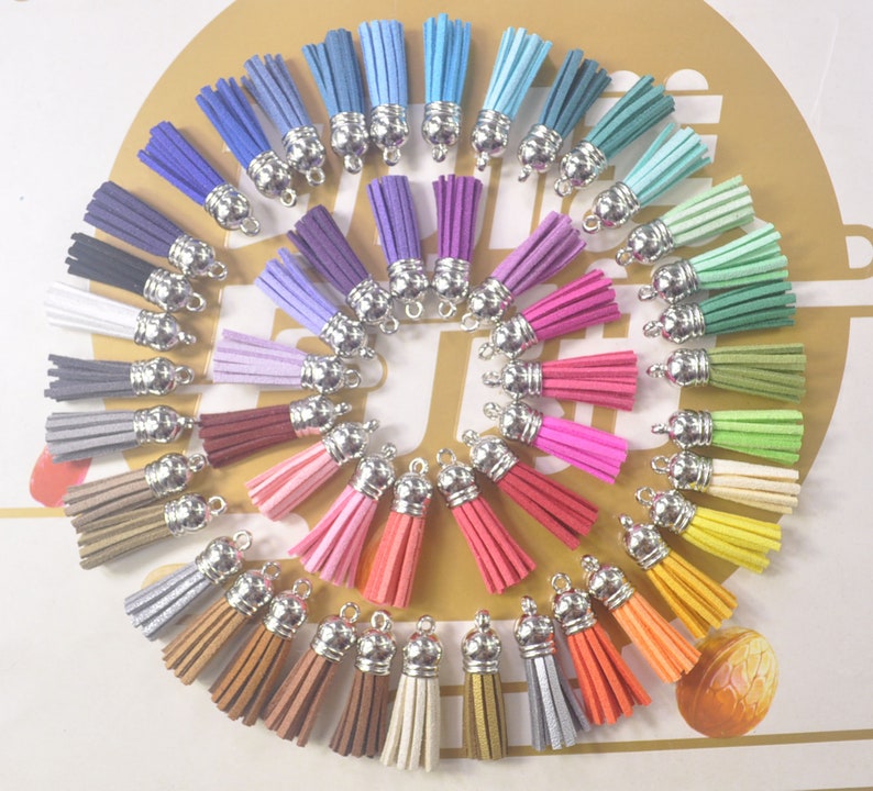 May include: A collection of colorful suede tassels in various shades, including blue, purple, pink, green, and orange. Each tassel has a silver-colored metal cap and a loop for attaching to jewelry or other crafts. The tassels are arranged in a circular pattern.