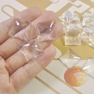 Puede incluir: Una mano sosteniendo varios adornos cuadrados de vidrio transparente. Los adornos son planos y tienen un ligero bisel alrededor de los bordes.