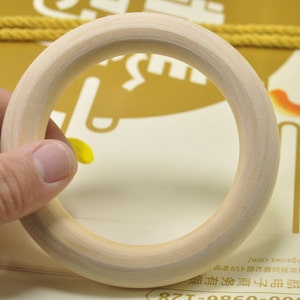 May include: A natural wood gymnastic ring, approximately 10 inches in diameter, with a smooth, light brown finish. The ring is held by a hand, showcasing its size and shape.