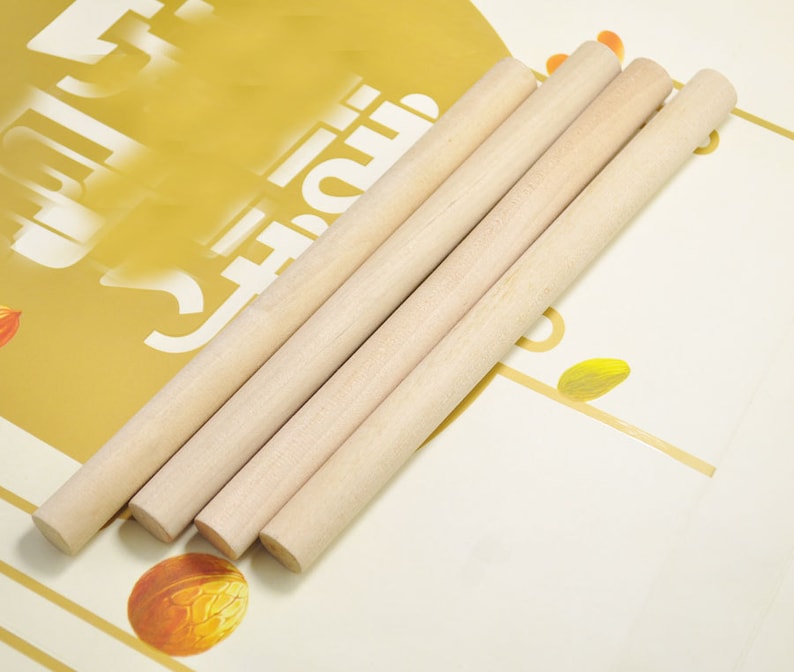 Puede incluir: Cuatro clavijas de madera natural, de aproximadamente 30 cm de largo, est&aacute;n dispuestas en una fila sobre un fondo blanco y dorado. Las clavijas son lisas y tienen un color marr&oacute;n claro.