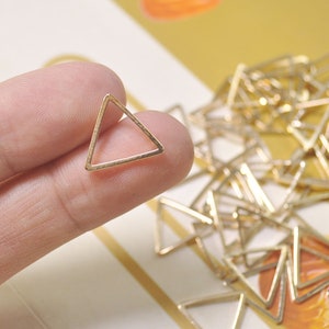 May include: A close-up of a gold triangle-shaped metal ring. The ring is being held by a finger. There are many more gold triangle rings in the background.