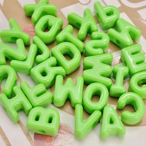 May include: A collection of bright lime green plastic alphabet beads, each featuring a molded letter of the alphabet with a smooth, glossy finish and a central hole for threading, scattered together in a random arrangement.