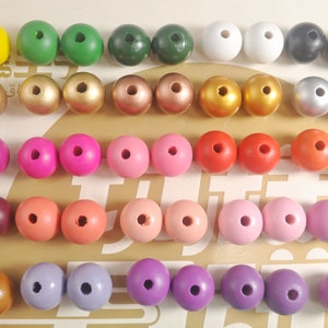 May include: A collection of 40 wooden beads in various colors, including yellow, green, white, black, gold, silver, pink, red, brown, and purple. The beads are arranged in rows on a light brown surface.