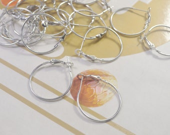 May include: A collection of silver hoop earrings. The earrings are round with a hinged closure. Several earrings are scattered on a light-coloured surface with gold and brown stripes.