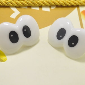 May include: Two white, cartoon-style eye-shaped embellishments with large black pupils. The glossy, plastic-like eyes are set against a light yellow background with a gold rope trim. These could be used for crafts or as decorative elements.