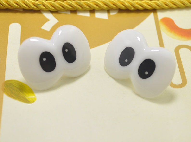 May include: Two white, cartoon-style eye-shaped embellishments with large black pupils. The glossy, plastic-like eyes are set against a light yellow background with a gold rope trim. These could be used for crafts or as decorative elements.