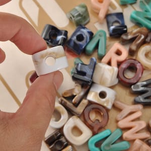 May include: A hand holding a rectangular, off-white bead with a circular hole. The bead has a marbled pattern with tan and white hues. Other letter-shaped beads in various colors and patterns are scattered in the background.