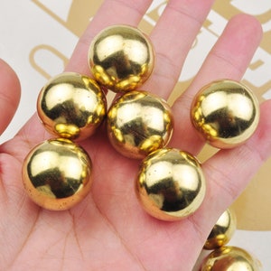 May include: A group of six shiny gold metal balls, likely made of brass, are arranged in a hand. The balls are smooth and reflective.