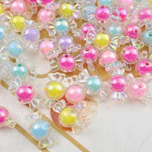 Puede incluir: Surtido de cuentas iridiscentes con forma de caramelo en rosa, azul, amarillo y transparente. Las cuentas son de plástico y tienen un acabado holográfico.
