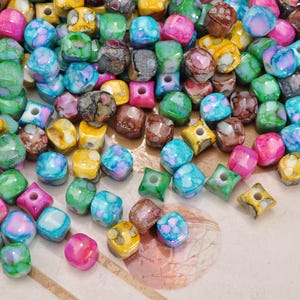 May include: A collection of colorful, mixed-shape beads in various shades of pink, blue, green, yellow, and brown. The beads have a marbled or speckled appearance, with some featuring square or star-like holes. These beads are suitable for jewelry making.