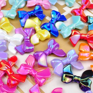 Op de afbeelding: Assortiment van regenboogkleurige, plastic charmes in de vorm van strikken in verschillende kleuren, waaronder roze, blauw, paars, geel, oranje en wit.