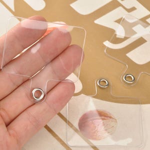 Può includere: Etichette rettangolari in plastica trasparente con un piccolo foro e un occhiello metallico. Le etichette sono tenute in mano, con uno sfondo sfocato. Le etichette sembrano essere per gioielli o artigianato.