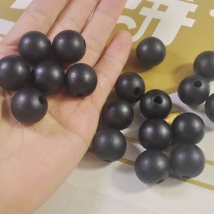 May include: A collection of black wooden beads, some are in a hand, others are scattered on a surface. The beads are round and have a smooth, matte finish.