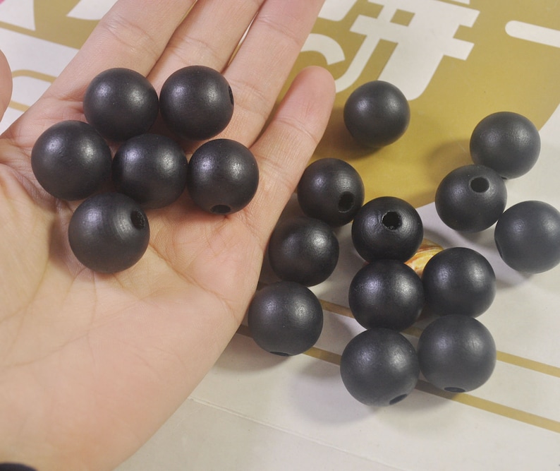 May include: A collection of black wooden beads, some are in a hand, others are scattered on a surface. The beads are round and have a smooth, matte finish.