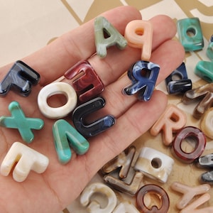 May include: A collection of colorful letter beads in various colors and patterns, including blue, red, green, and white. The beads are in different fonts and sizes, perfect for jewelry making or crafting projects.