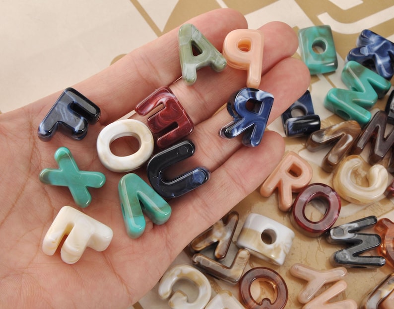May include: A collection of colorful letter beads in various colors and patterns, including blue, red, green, and white. The beads are in different fonts and sizes, perfect for jewelry making or crafting projects.