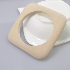 May include: A light-colored, square wooden bracelet with rounded corners. The bracelet is displayed on a white ceramic dish, set against a gray background. The bracelet's design is simple and modern, showcasing the natural wood grain.