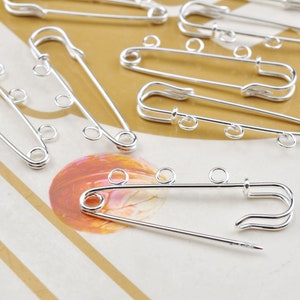 May include: A set of silver safety pins with multiple loops. The pins are arranged on a striped surface.
