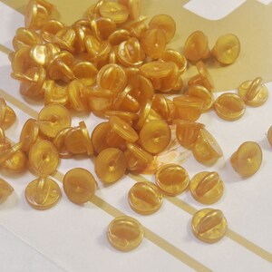 Könnte beinhalten: Ein großer Haufen goldfarbener Sicherheitsnadelrücken. Die Rücken sind rund und haben einen kleinen Stift, der aus der Mitte herausragt.