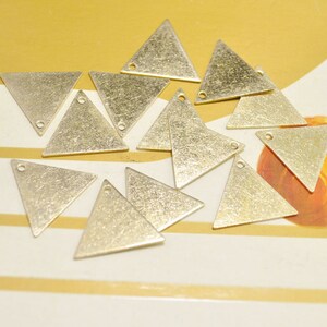May include: A group of twelve gold-toned triangle-shaped metal charms with a hole at the top. The charms have a textured surface and are scattered on a striped surface.