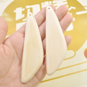 May include: Two light-colored wooden pendants with a smooth, curved design. Each pendant has a small hole at the top for stringing. The pendants are held in a hand, showcasing their natural wood grain and shape.